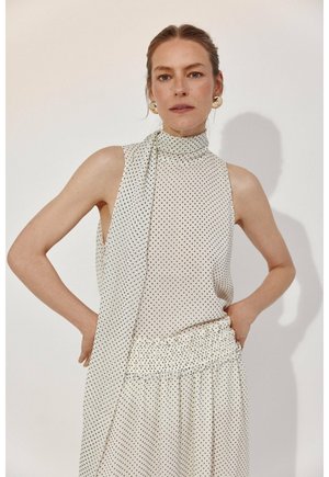 Femme portant une robe blanche sans manches à pois avec un col haut et une taille froncée, debout les mains sur les hanches devant un fond uni.