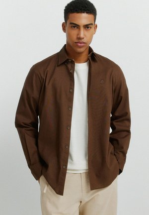 Młody mężczyzna w brązowej koszuli na guziki, nałożonej na białą koszulkę, oraz beżowych spodniach, stojący na jasnym, jednolitym tle.