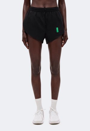 Personne portant un short de sport noir avec une étiquette verte, des chaussettes blanches et des baskets blanches, debout devant un fond uni.