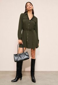 Rochie kaki cu nasturi, mâneci lungi, talie strânsă și buzunare. Asortată cu cizme negre până la genunchi și o geantă neagră.