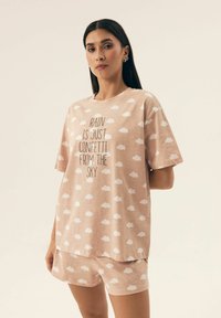Jasnoróżowy zestaw odzieży domowej składający się z T-shirta z białymi wzorami chmur i czarnym napisem. Szorty mają ten sam wzór chmur. Luźny krój.