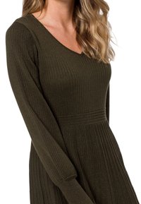 Robe vert olive foncé avec une texture côtelée, manches longues, décolleté en V et corsage ajusté qui s'évase à la taille, avec un subtil éclat.