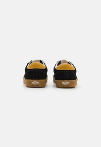 Zapatillas de ante negro con detalles en amarillo, suela de goma y un distintivo logo de Vans. La vista trasera resalta la costura y la forma.