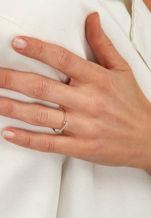 Silberring mit einem einzelnen Diamanten. Glatter, dünner Ring mit einem subtilen Twist-Design. Hand ruht auf weißem, strukturiertem Stoff.