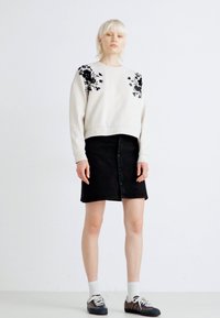 Kremna pot sweatshirt z black cvetličnim vezenjem, v kombinaciji z črno srajčasto krilo. Manekenka nosi bele nogavice in črne tekaške copate.