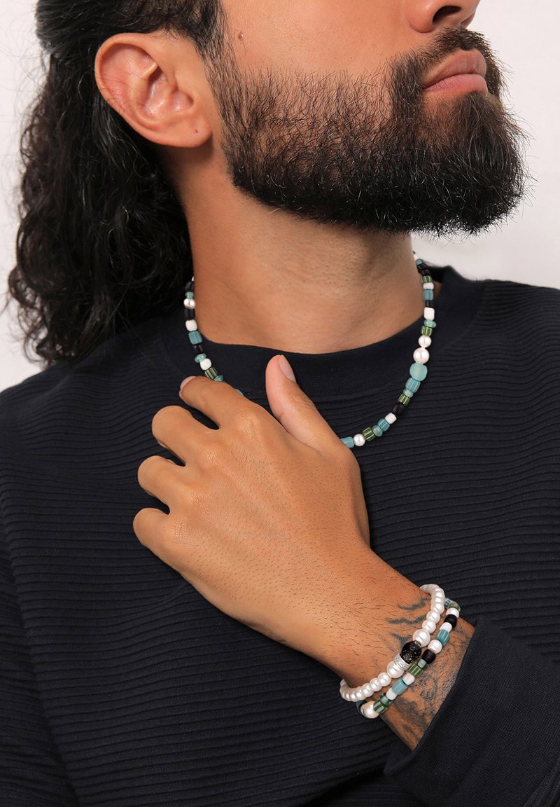 Skægget mand med langt hår iført en sort ribbet skjorte, en perlekæde og tre perlearmbånd på sit tatoverede håndled.