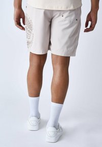 Pantalones cortos de color beige claro en una tela ligera, con un gran estampado gráfico en la pierna izquierda y dos bolsillos traseros con cierres a presión.