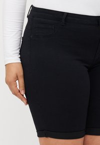 Musta denimhousut, joissa on vartalonmyötäinen istuvuus, keskikorkea vyötärö, käännetty lahje ja hillittyjä ompelu- ja yksityiskohtia.