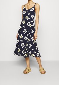 Blumenkleid in Marineblau mit weißen und gelben Blumen. V-Ausschnitt, schmale Träger, ausgestellt am Saum. Kombiniert mit beigen Sandalen für einen lässigen Look.
