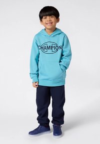 Χαρούμενος αγόρι που φοράει μια ανοιχτό μπλε φούτερ Champion, σκούρα μπλε φόρμες και μπλε αθλητικά παπούτσια, στέκεται μπροστά σε ένα απλό λευκό φόντο.