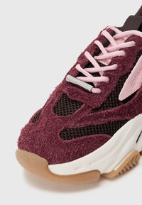 Zapatilla burdeos y negra con textura aterciopelada, detalles de malla, cordones rosas y una entresuela blanca. Presenta un detalle de logo plateado en el lateral.