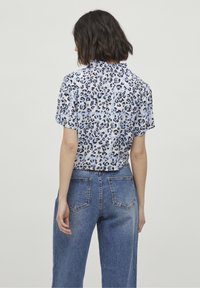 Donna con capelli corti e scuri indossa una camicia a maniche corte con stampa leopardata infilata in jeans blu a vita alta, in piedi di fronte a uno sfondo neutro.