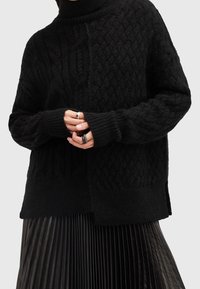 Pull noir en maille avec motifs texturés, col haut, épaules tombantes et poignets côtelés. Porté avec une jupe noire plissée.