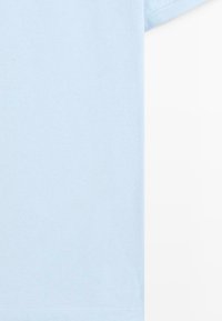 Hellblaues kurzärmeliges Poloshirt in Nahaufnahme, zeigt die Stoffstruktur und einen Teil des rechten Ärmels vor weißem Hintergrund.