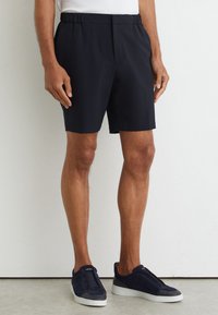 Man wearing black elastic waist shorts and navy blue sneakers standing against a white textured wall.