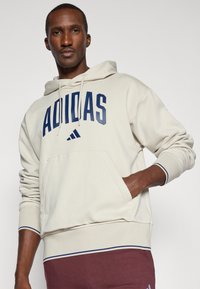 Kremna kapuća sa velikim mornarskim "ADIDAS" logom, sa kengurujim džepom i rebrastim manžetama sa tamnoplavim akcentima.