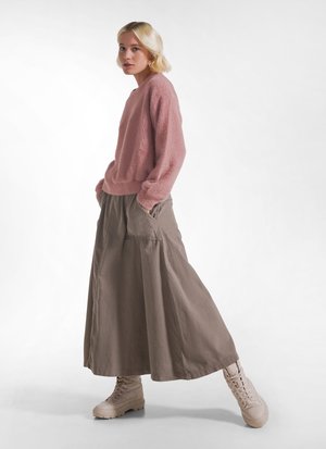 Kvinde med kort blondt hår iført en lyserød sweater, lang grå nederdel med lommer og beige snørestøvler, går med hænderne i lommerne.