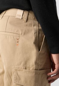 Beige cargo pants with a straight cut, featuring a back pocket, three orange embroidered accents, and a branded waistband label.