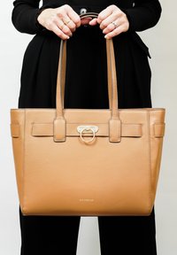 Sac fourre-tout en cuir tan avec poignées supérieures doubles, présentant une sangle centrale avec un accent en métal doré en forme de cercle et des détails cousus.