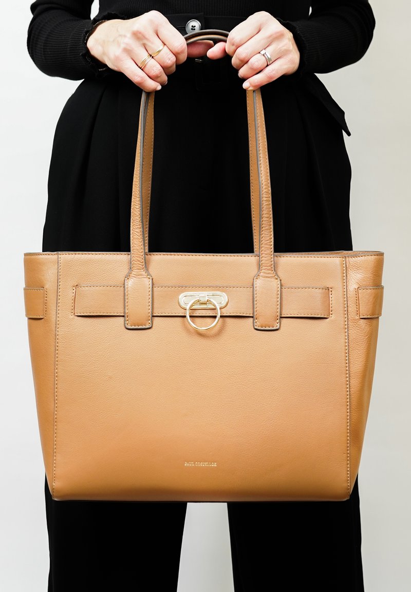 Sac fourre-tout en cuir tan avec poignées supérieures doubles, présentant une sangle centrale avec un accent en métal doré en forme de cercle et des détails cousus.