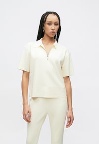 Κρεμ τοπ κοντομάνικο με γιακά μισής φερμουάρ, κατασκευασμένο από λείο, υφασμένο ύφασμα, με ραφές στους ώμους που πέφτουν χαμηλά και χαλαρή εφαρμογή.