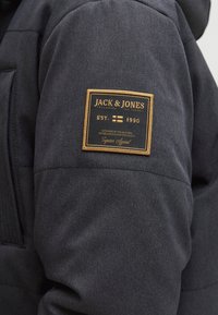 Ciemnoszara kurtka z materiałową naszywką z napisem "JACK & JONES" w kolorze złotym, zawierającą datę założenia i szczegółowe przeszycia dodające tekstury.