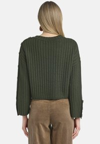 Ciemnozielony dziany sweter o prążkowanej fakturze, o skróconej długości, z guzikami na rękawach, zestawiony z jasnobrązowymi sztruksowymi spodniami.