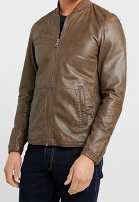 Giubbotto bomber in pelle marrone con chiusura zip, colletto a costine, due tasche laterali e una texture liscia. Silhouette aderente con una leggera lucentezza.