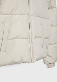 Beige puffjacka med horisontell sömnad, dragkedjestängning och sidofickor. Tillverkad av glänsande, slät tyg med en mjuk textur.