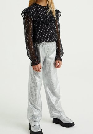 Dziecko w przezroczystej czarnej bluzce z falbankami i wzorzystymi kropkami, błyszczących srebrzystych spodniach o szerokich nogawkach oraz masywnych srebrnych butach na jednolitym tle.