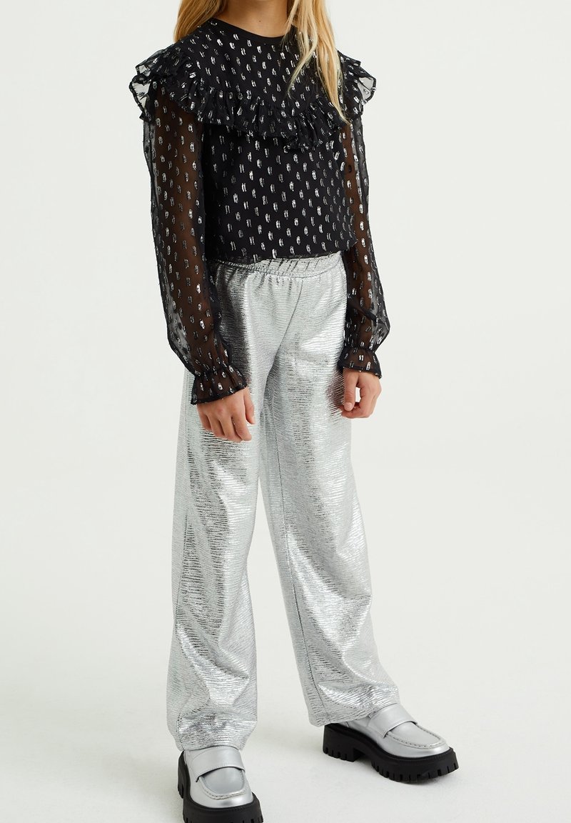 Enfant portant un chemisier noir transparent à volants avec des points à motifs, un pantalon large argenté brillant et des chaussures épaisses argentées sur un fond uni.