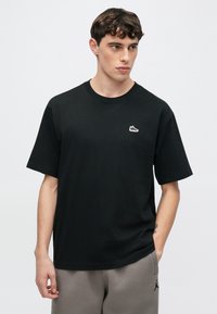 Μαύρο βαμβακερό μπλουζάκι με γραφικό λογότυπο παπουτσιού στο αριστερό στήθος, κοντά μανίκια και στρογγυλή λαιμόκοψη, συνδυασμένο με γκρι φόρμες.