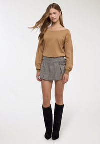 Maglione marrone scollato abbinato a una gonna a pieghe a quadri. Stivali neri che arrivano al ginocchio completano l'outfit. Texture morbide e motivi contrastanti sono visibili.