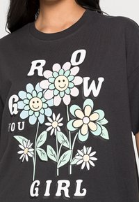 Černé bavlněné tričko s květinovou grafikou, na které jsou usmívající se tváře, pastelově barevné květiny a text "GROW YOU GIRL" tučnými bílými písmeny.