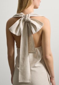 Beige Leinenkleid mit einem rückenfreien Design, das mit einer großen Schleife und langen, herabhängenden Bändern am Hals gebunden ist und eine strukturierte Oberfläche zeigt.