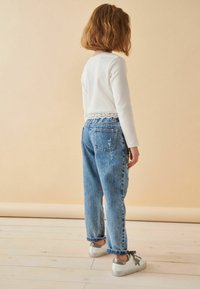 Camisa blanca de manga larga con dobladillo de encaje, combinada con jeans de denim azul desgastados y zapatillas grises con detalles plateados.