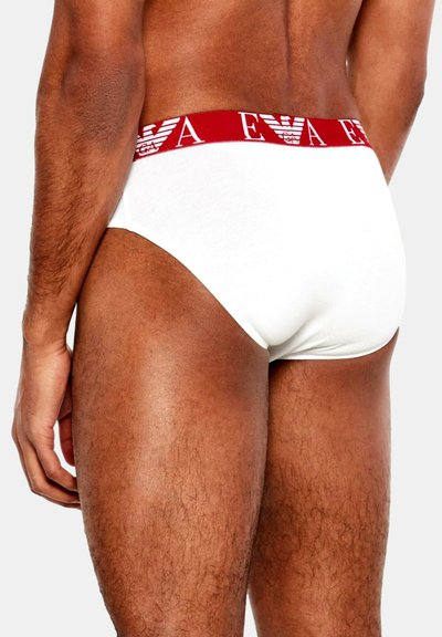 Białe bawełniane majtki z czerwonym elastycznym paskiem w pasie ozdobionym powtarzającym się wzorem logo. Dopasowanie jest przylegające, a tkanina wygląda na gładką.
