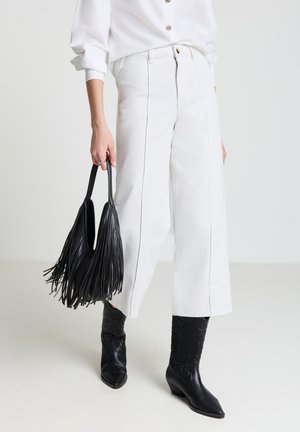 Pantalon large blanc en denim à taille haute, associé à un sac à main noir à franges et des bottines cowboy noires à bout pointu.