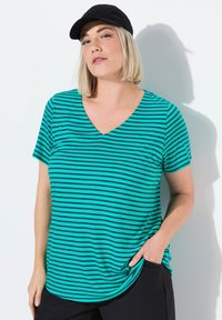 Top de manga corta en verde azulado con rayas horizontales negras y escote en V. Confeccionado en una tela suave, tiene un corte relajado y bolsillos laterales. Se lleva una gorra negra.