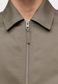 Κοντινό πλάνο του μπροστινού μέρους ενός χακί μπουφάν που δείχνει ένα μεταλλικό φερμουάρ μερικώς κλειστό κάτω από ένα κλασικό γιακά σε ανοιχτόχρωμο δέρμα.