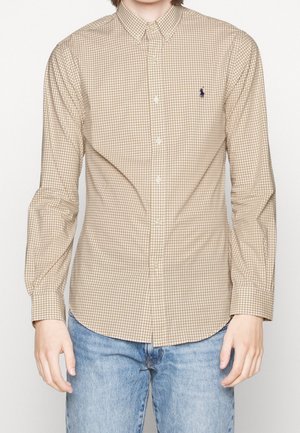 Bruine en beige geruite blouse met kraag, lange mouwen en een geborduurd logo op de borst, gecombineerd met blauwe jeans.