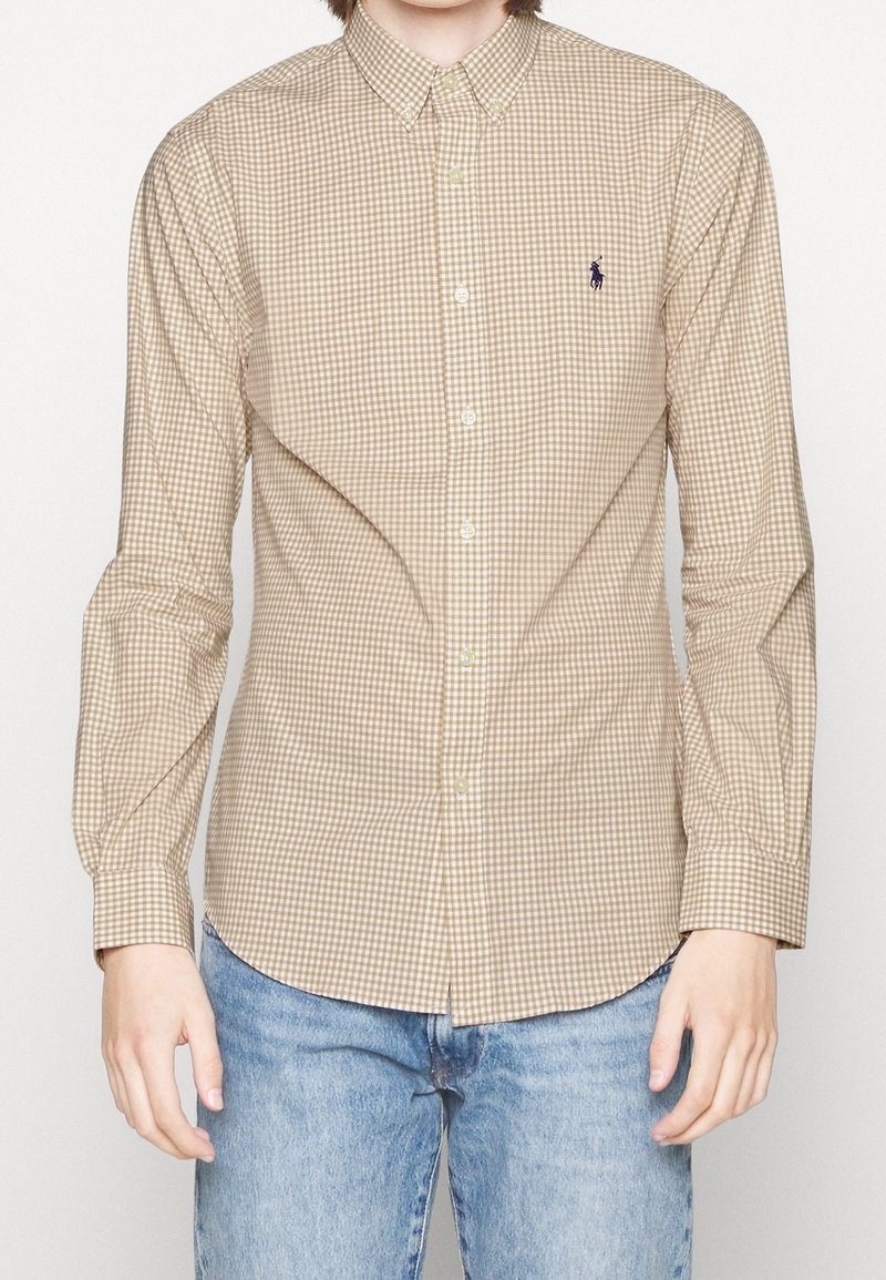 Bruine en beige geruite blouse met kraag, lange mouwen en een geborduurd logo op de borst, gecombineerd met blauwe jeans.