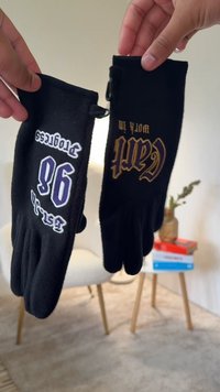 Černé fleecové rukavice zdobí bílé a zlaté nápisy. Levá rukavice nese fialové číslo "96" a bílý nápis "Protects"; pravá rukavice má zlatý nápis "Craft Worth It".
