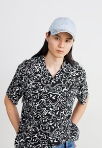 Maglietta nera a maniche corte con motivi astratti bianchi, abbinata a un cappellino blu chiaro con un logo e dettagli floreali. Jeans di denim.