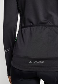 Black long-sleeve athletic top featuring a textured fabric, reflective accents, and a rear pocket with a green label. Logo at the waist.
