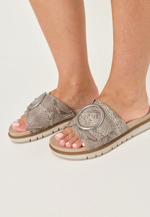 Pieds portant des sandales à enfiler beige imprimé serpent avec une grande boucle métallique circulaire et une semelle blanche épaisse, sur fond blanc.