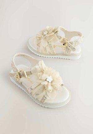 Beežid sandaalid võltslilledega, sädelevate rihmade ja tekstuurse valge tallaga. Reguleeritav pandla turvaliseks sobivuseks.