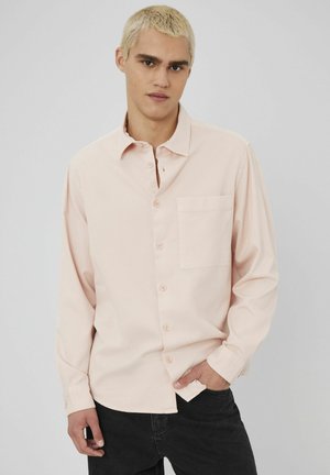 Chemise boutonnée à manches longues de couleur rose clair, avec un col, une poche poitrine unique et une fermeture à boutons sur le devant, portée avec un jean foncé.