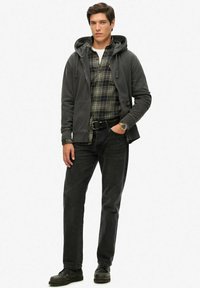 Joven con camisa de cuadros, sudadera negra, jeans negros, reloj y zapatos negros, de pie con una mano en el bolsillo sobre un fondo blanco.