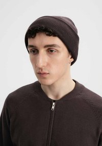 Bruine gebreide beanie met ribbelstructuur. Model draagt een bruine sweater met rits en een gestructureerde halslijn, met een zilveren ritsdetail. Neutrale achtergrond.